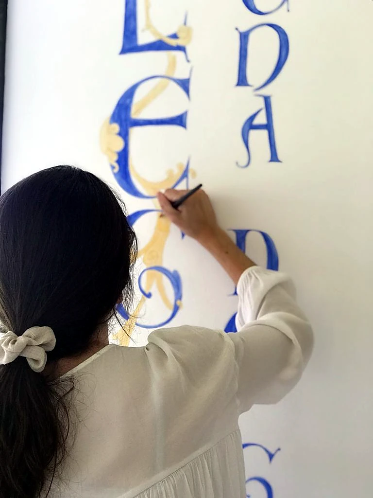 Lidia pintando versales azules y doradas sobre un muro
